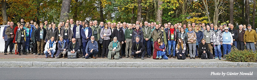 The conference participants line of for a photo in front of a wooded area. Text: Photo by Gunter Nowold