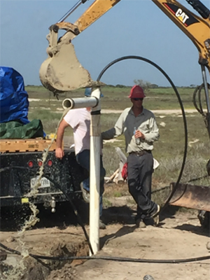 A work crew installs a well to create a new fresh water pond. A large, white pipe is coming out of the ground, and yellow construction equipment are in the background.
